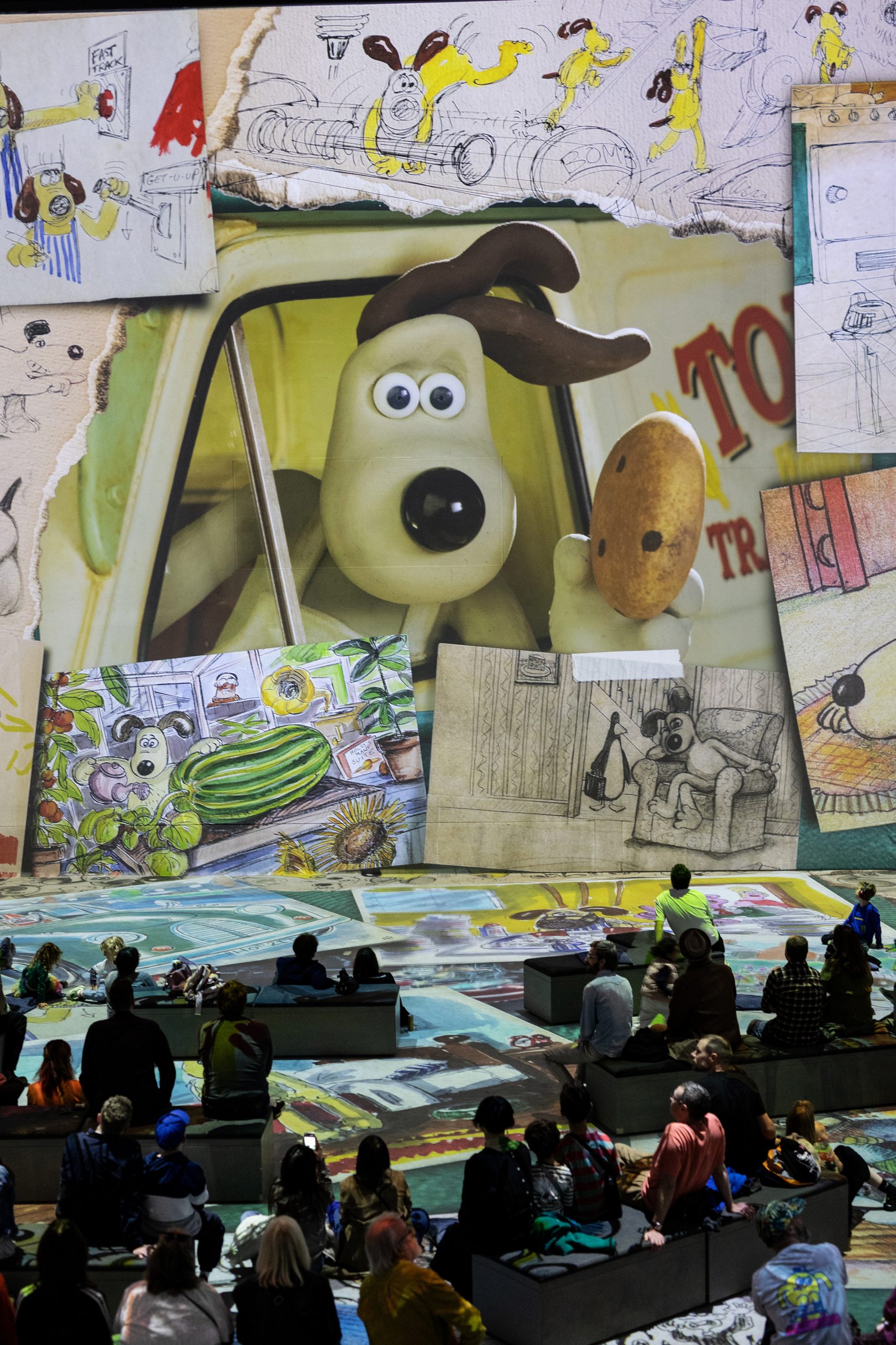 Gromit and original development sketches projected above audiences seated on a floor of artwork