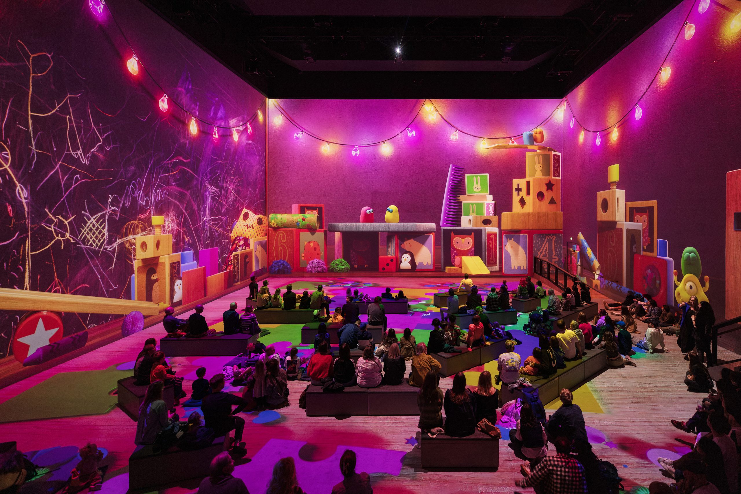Visitors sit among giant projected toy blocks and string lights in the Aardman immersive experience at Lightroom