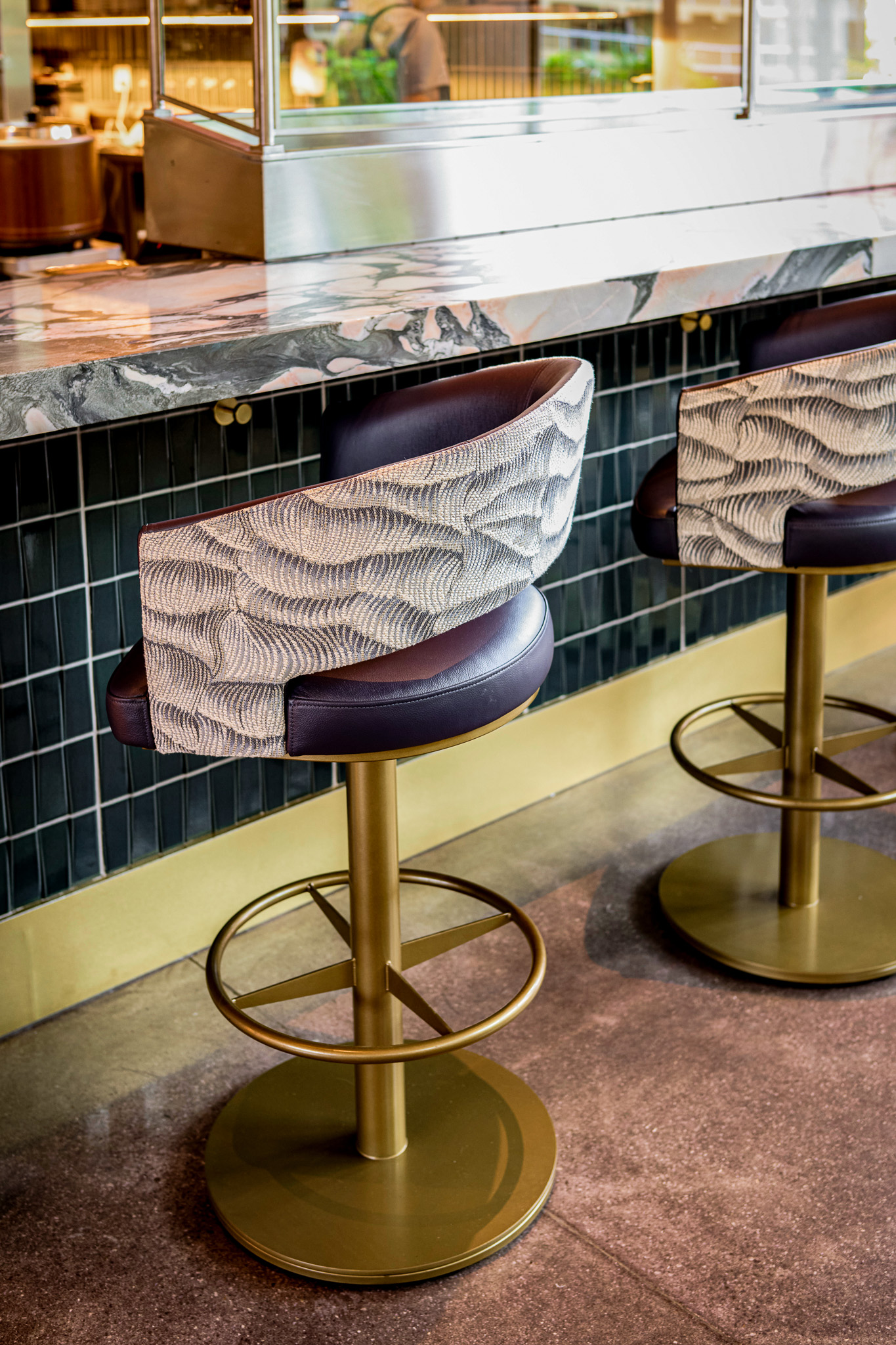 Upholstered bar stools with brass bases at a marble counter with dark green tile