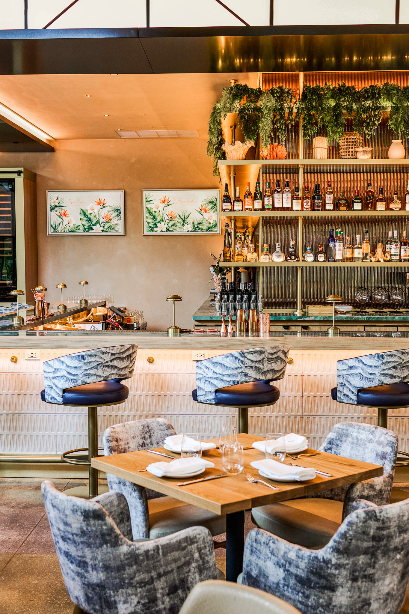 Bar seating and dining tables in front of a back bar with shelving, greenery and artwork