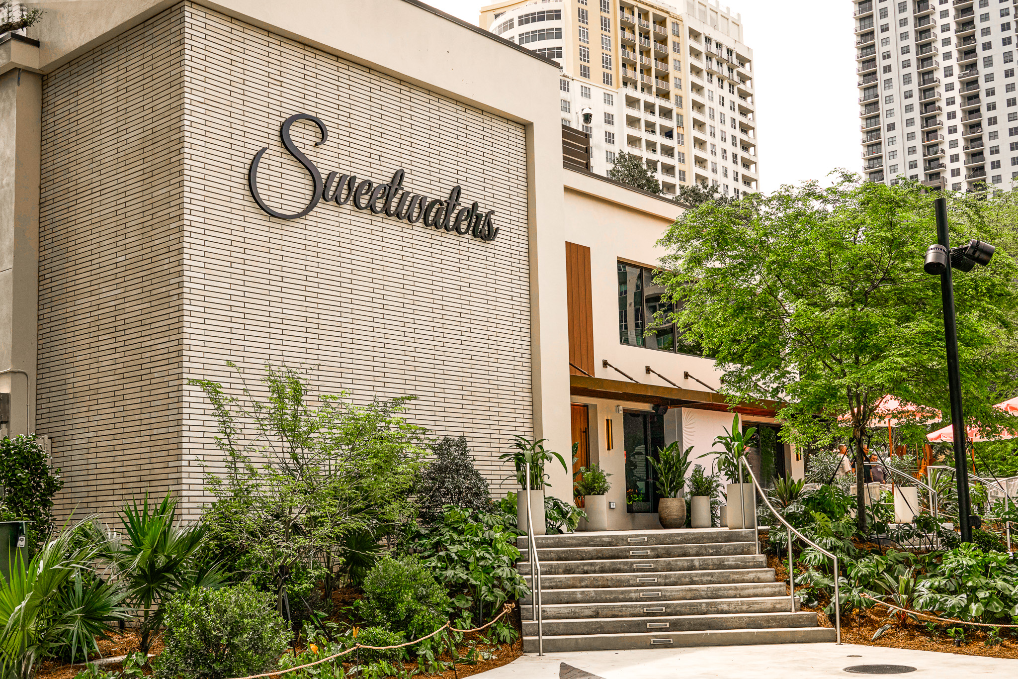 The white brick facade and signage of Sweetwaters, a waterfront restaurant in Huizenga Park, above planted steps